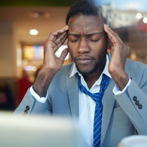 Businessman rubbing his temples suffering from headache while sitting at counter in coffee shop, shot behind glass window