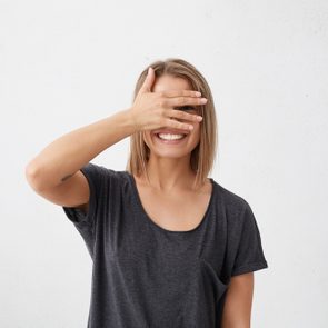 Merry woman having shy look peeking through her fingers demonstrating her even white teeth. Embarrassed young cute female with bob hairdo covering her face with hand looking confusedly smiling broadly