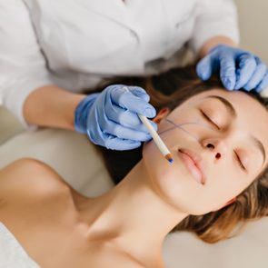 Portrait woman with brunette hair at preparation to rejuvenation, cosmetology operation in beauty salon.  Hands in blue gloves drawing on face, botox, beauty