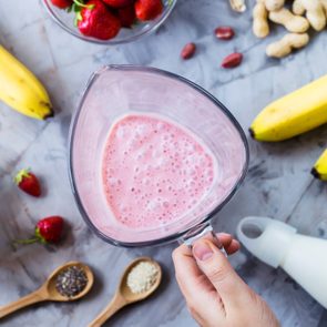 Ingredients for making strawberry banana smoothies on a gray table next to a glass bowl of blendet. Cooking a healthy breakfast concept. TOp view, flat lay