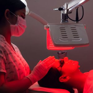 Young Woman Having Red Led Light Facial Therapy Treatment In Beauty Salon. Beautician Wearing Face Mask Maintaining Safety Procedures During Appointment. Beauty, New Normal And Wellness