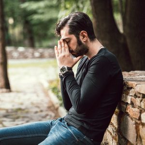 Young adult experience everyday stress sitting on bench. Holding his head with hands. Stylish clothing with bracelet and expensive watch.