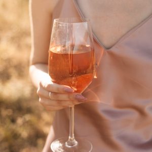 Young woman with long black hair wearing silk pink glass holding glass of rose wine posing outdoors in summer countryside